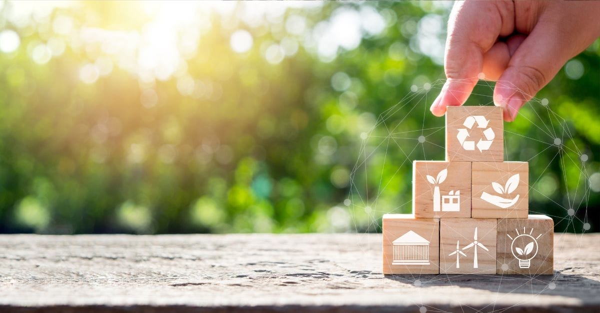 Sunshining through green tree leaves in the background. On a table, 5 wooden blocks put into a pyramid shape, each block representing an icon relating to the environment.