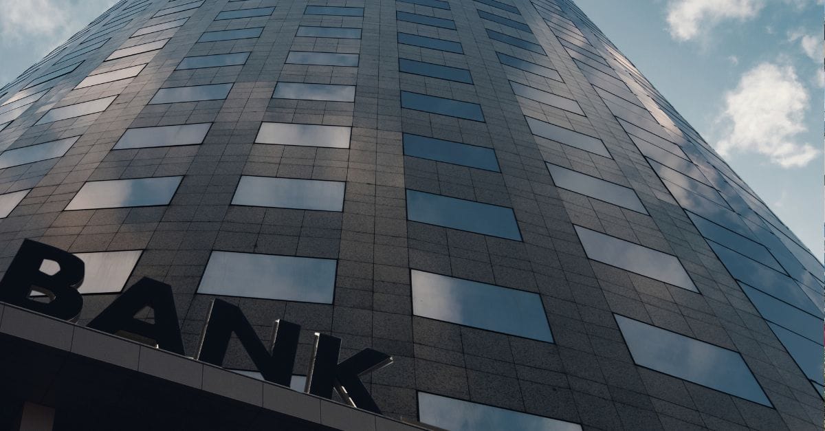 Outside view of a bank in concrete and glass windows. The word Bank is posed at the bottom of the building.